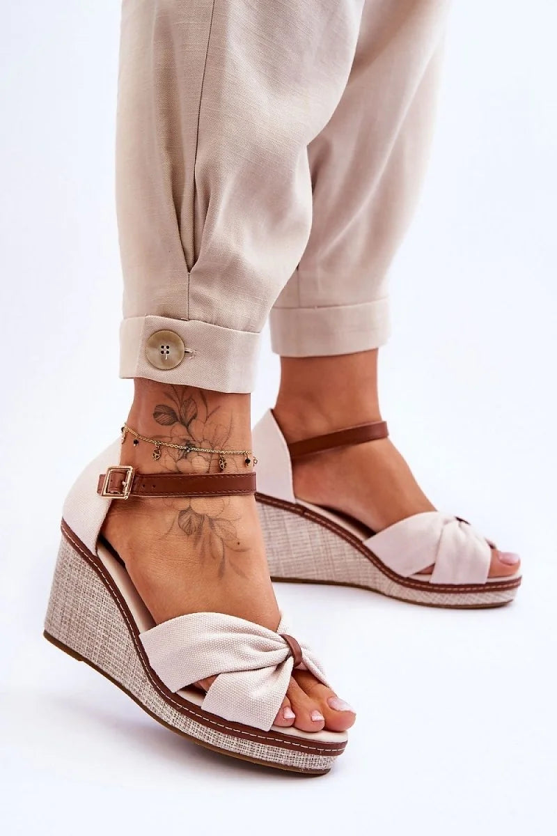 Beige fabric wedge sandals with brown straps and a bow detail on a model's foot.