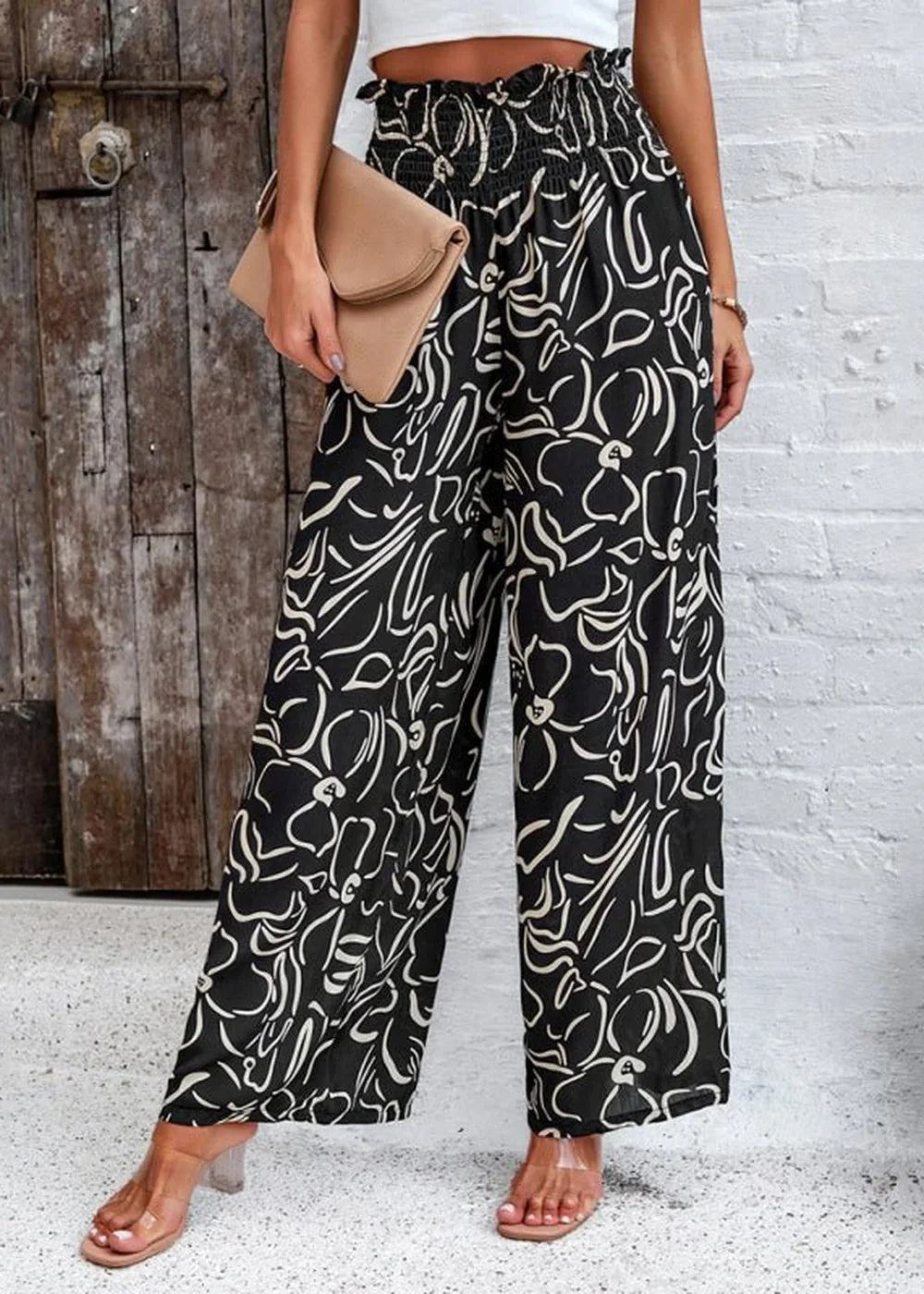 Wide-leg black pants with a white abstract floral print and a smocked waistband, worn with clear heeled sandals.