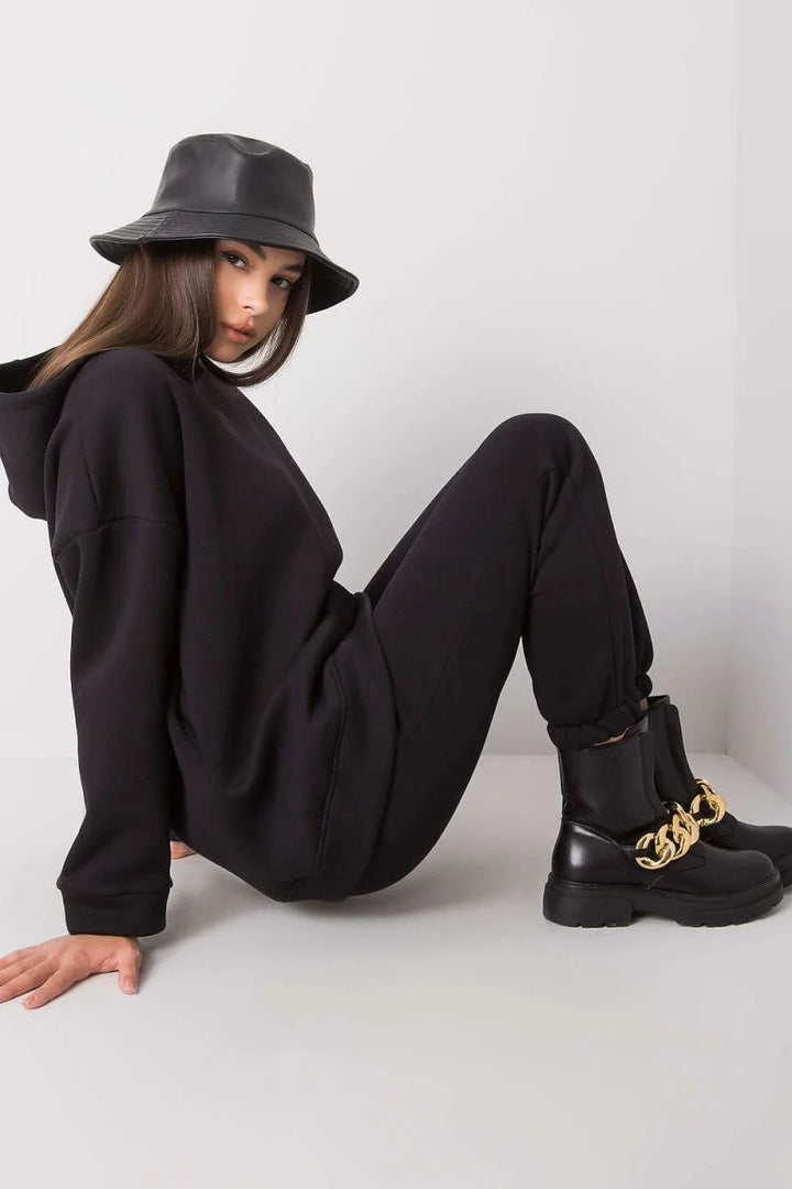 Black sweatpants with gold chain detail on boots, worn by a model in a black bucket hat and hoodie.