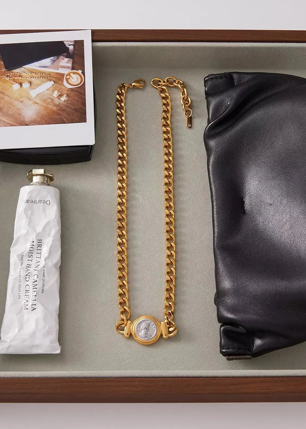 Gold-plated chain necklace with a coin pendant, next to a white hand cream tube and black leather gloves.