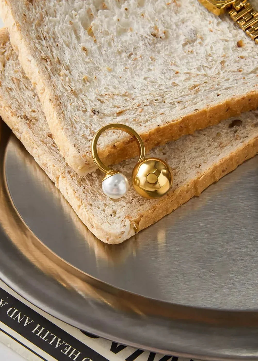 Gold ring with pearl and sphere charm on toast slices on a silver tray.