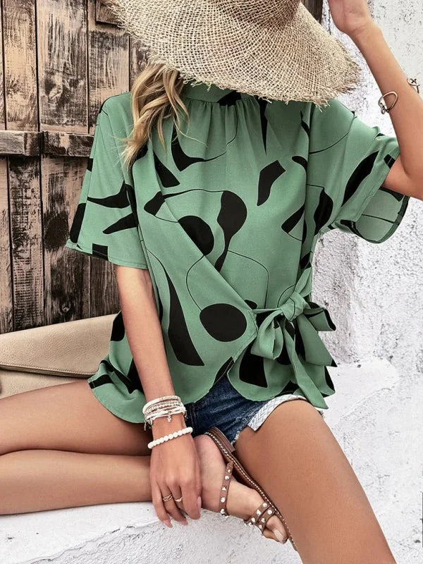 Green short-sleeve top with black abstract print, tie waist, worn with denim shorts and sandals.