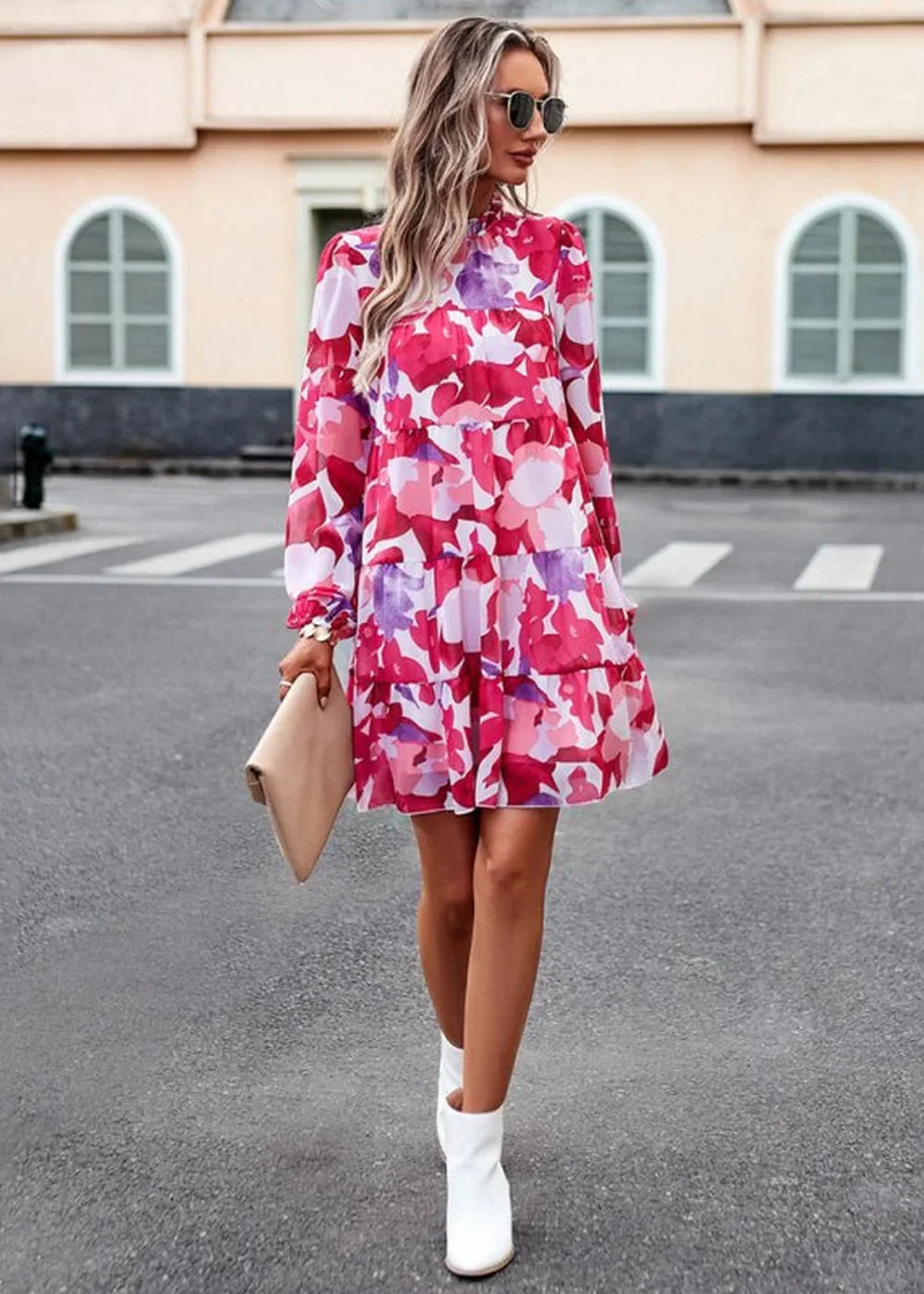 A woman wears a vibrant floral print mini dress with long sleeves and a tiered skirt, holding a clutch.