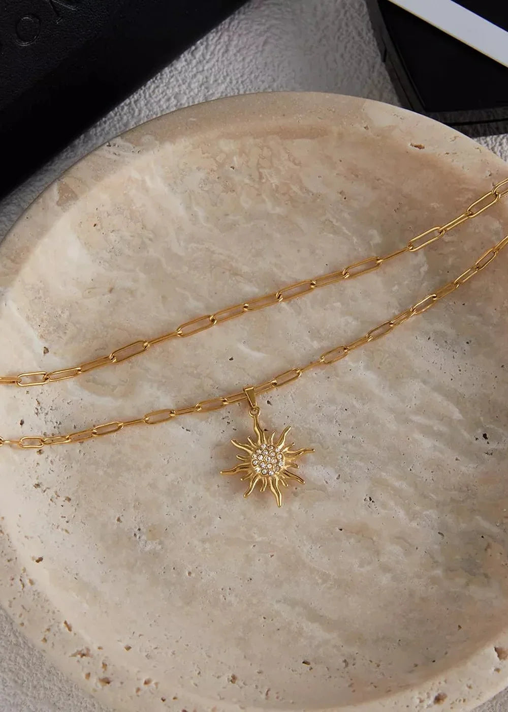 Gold layered necklace with a sparkling sun pendant on a textured stone surface.