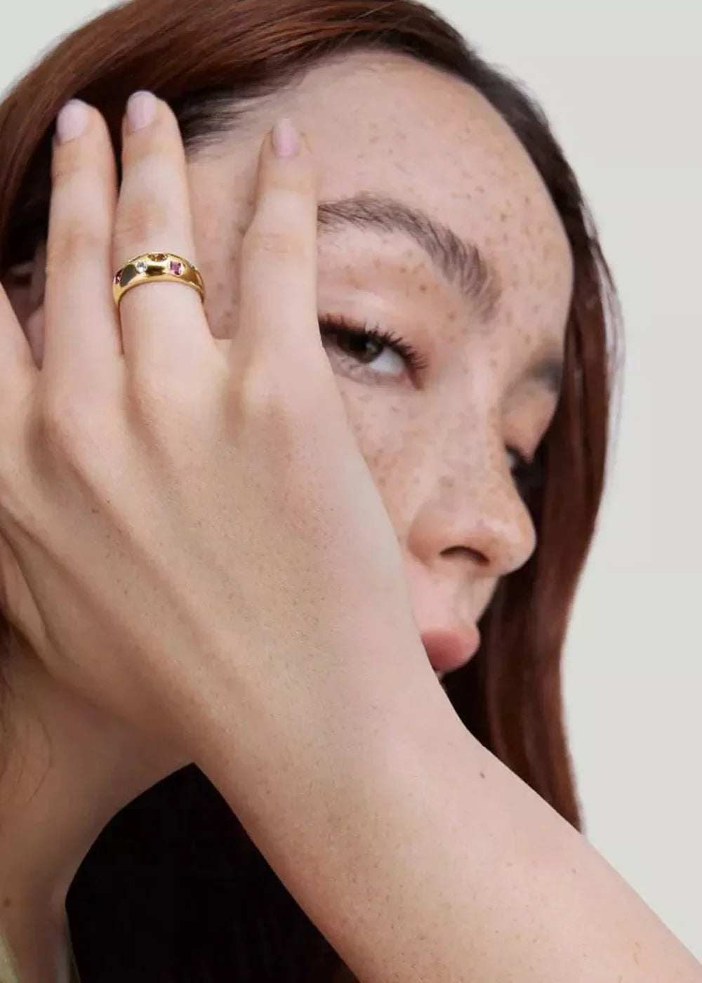 Gold ring with colorful stones on a hand, close-up of a woman's face with freckles.