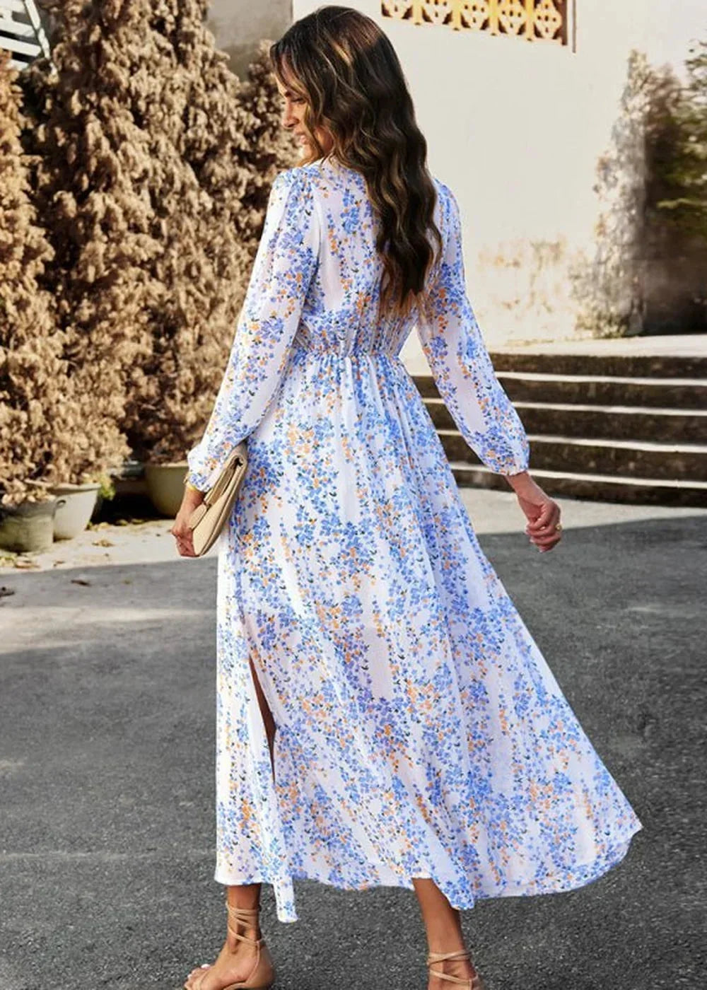 Woman in a flowing white maxi dress with a blue and orange floral pattern, featuring long sleeves and a thigh-high slit.