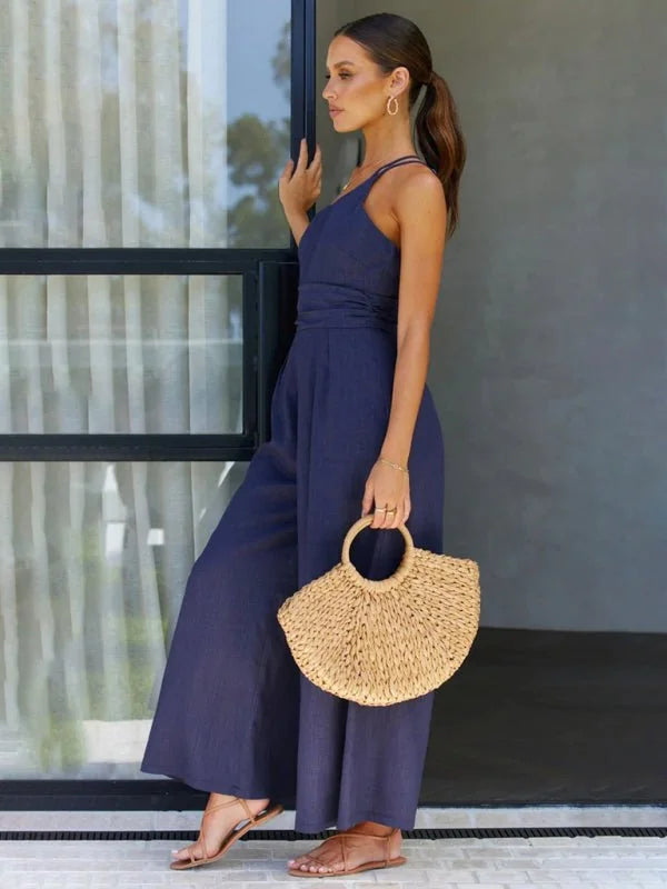 Woman in a navy blue wide-leg jumpsuit and straw bag, standing by a window.