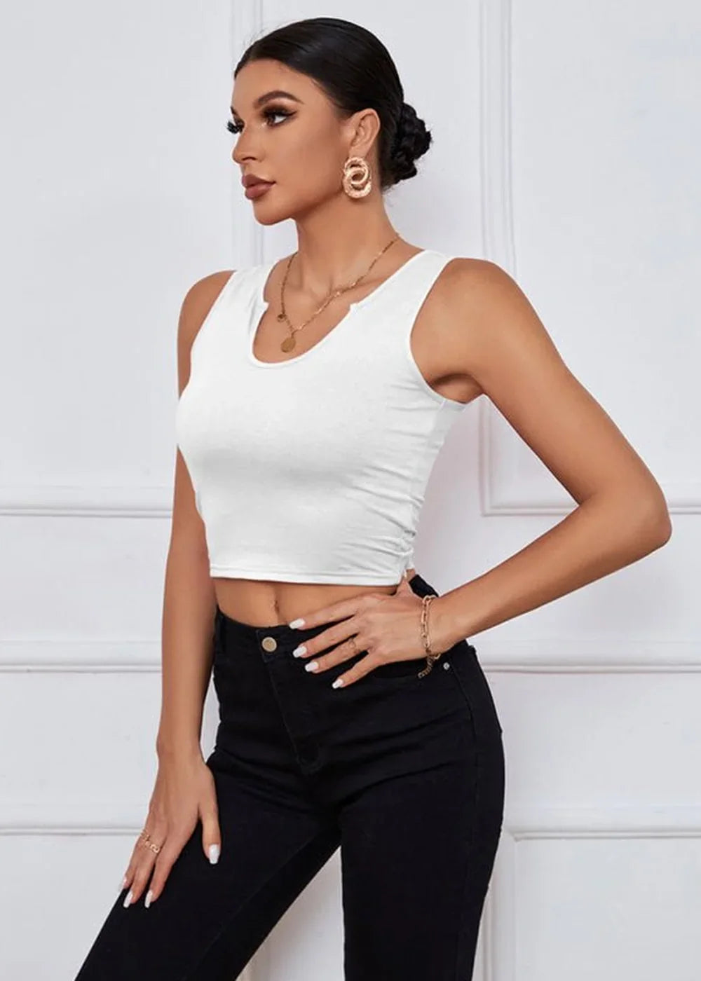 Woman in a white sleeveless crop top and black jeans, accessorized with gold jewelry.
