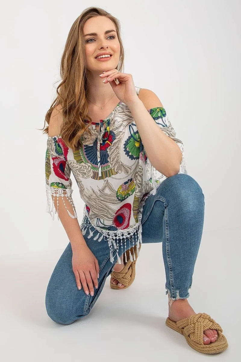 White off-the-shoulder top with colorful floral print, fringe trim, and blue distressed jeans.