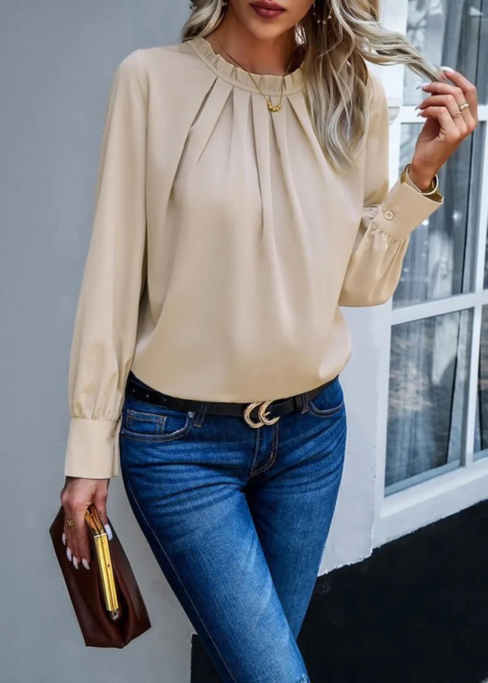Beige pleated long-sleeve blouse with ruffled neckline tucked into blue denim jeans and a black belt with a gold buckle.