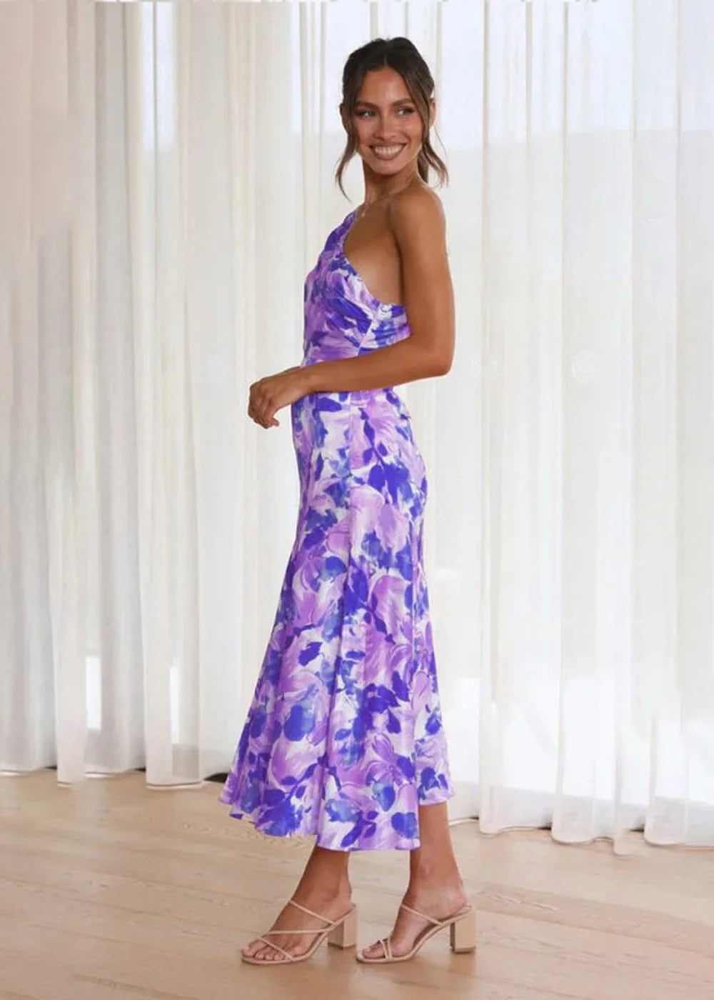 Woman in a purple and blue floral print one-shoulder midi dress with nude heeled sandals.