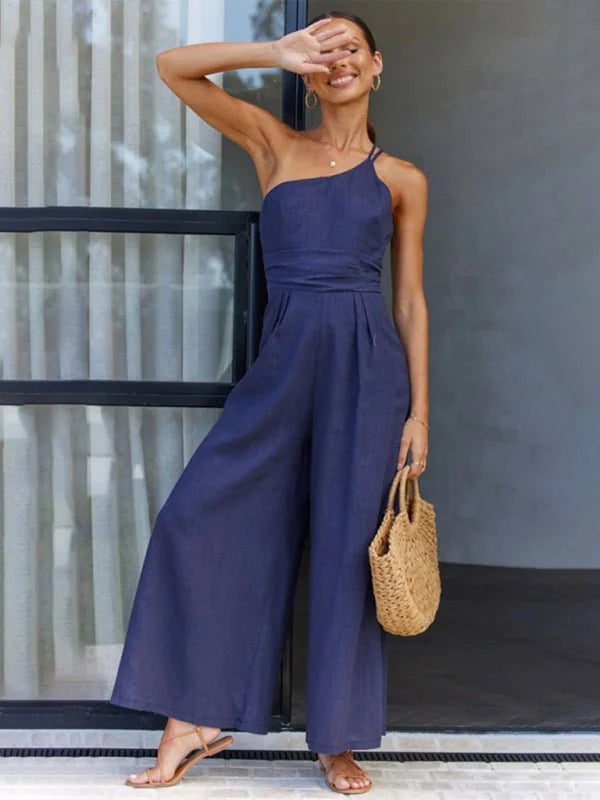 Navy blue one-shoulder wide-leg jumpsuit with a straw bag, shown on a model.