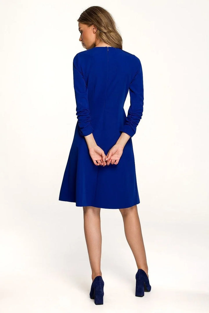 Back view of a royal blue dress with long ruched sleeves and a flared skirt, worn by a model with navy heels.