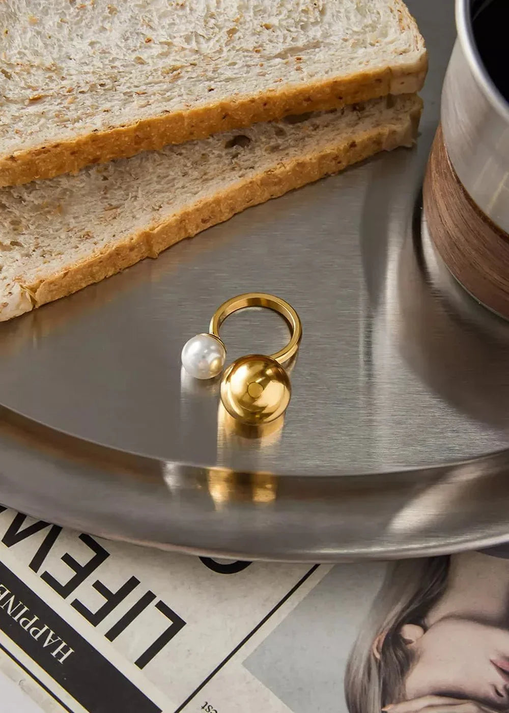 Gold ring with a pearl and a shiny gold sphere on a brushed metal tray with toast.