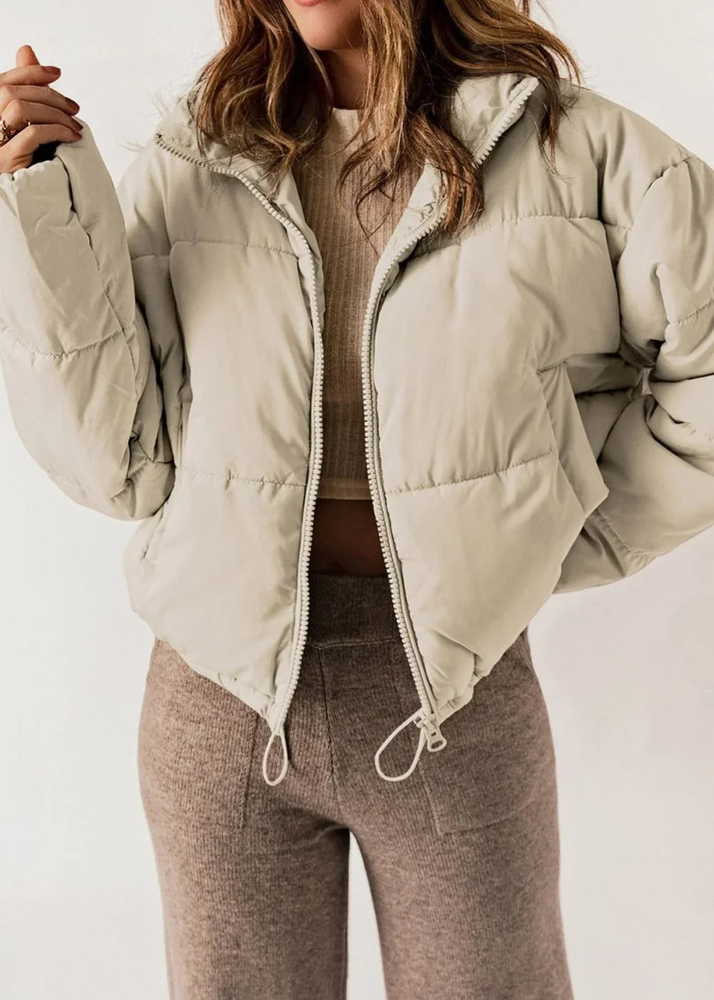 Light beige puffer jacket with a zippered front, worn by a model with brown ribbed pants.