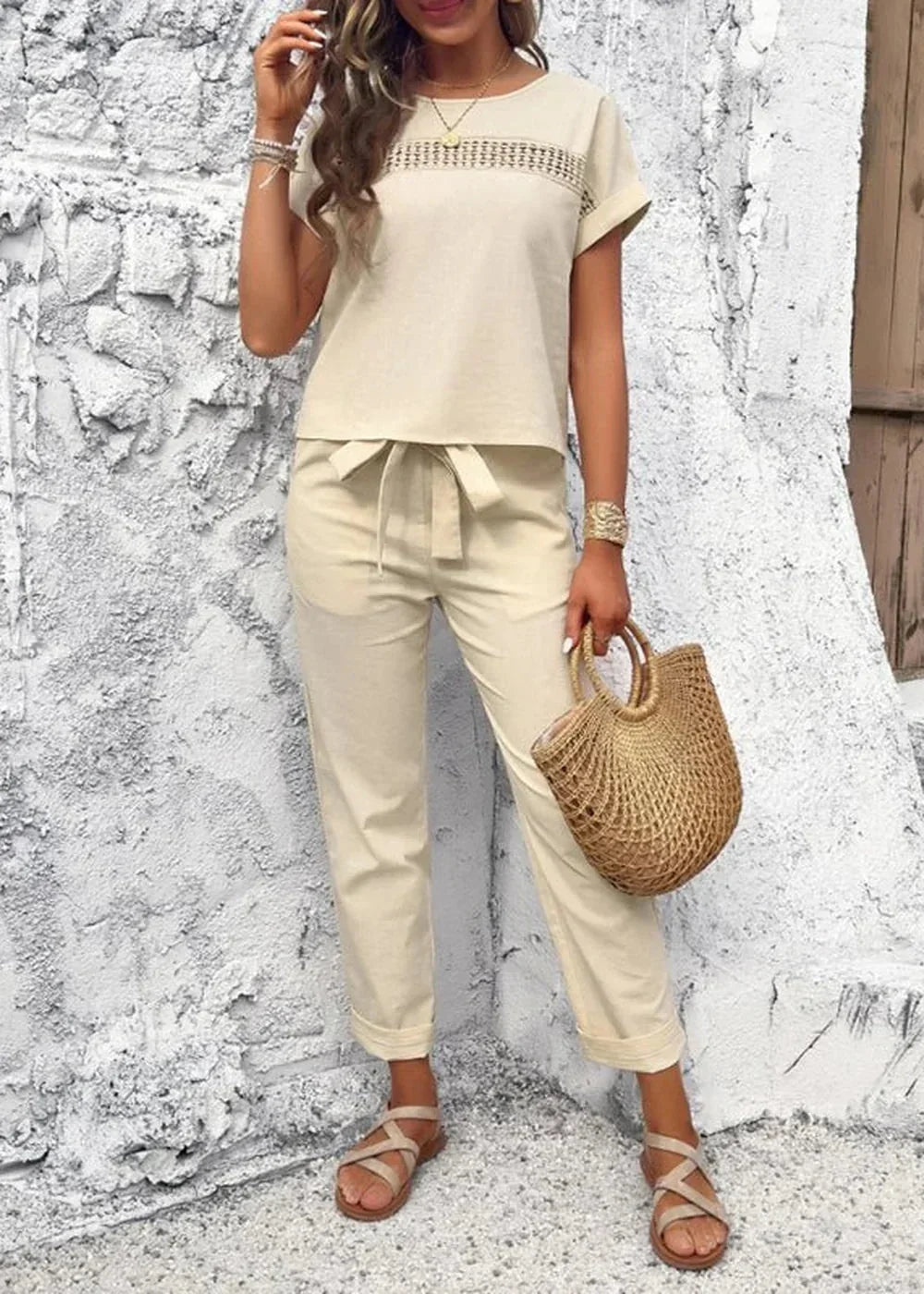 Light beige linen-blend pant suit with tie waist pants and crochet detail top, worn by a model with straw bag and sandals.