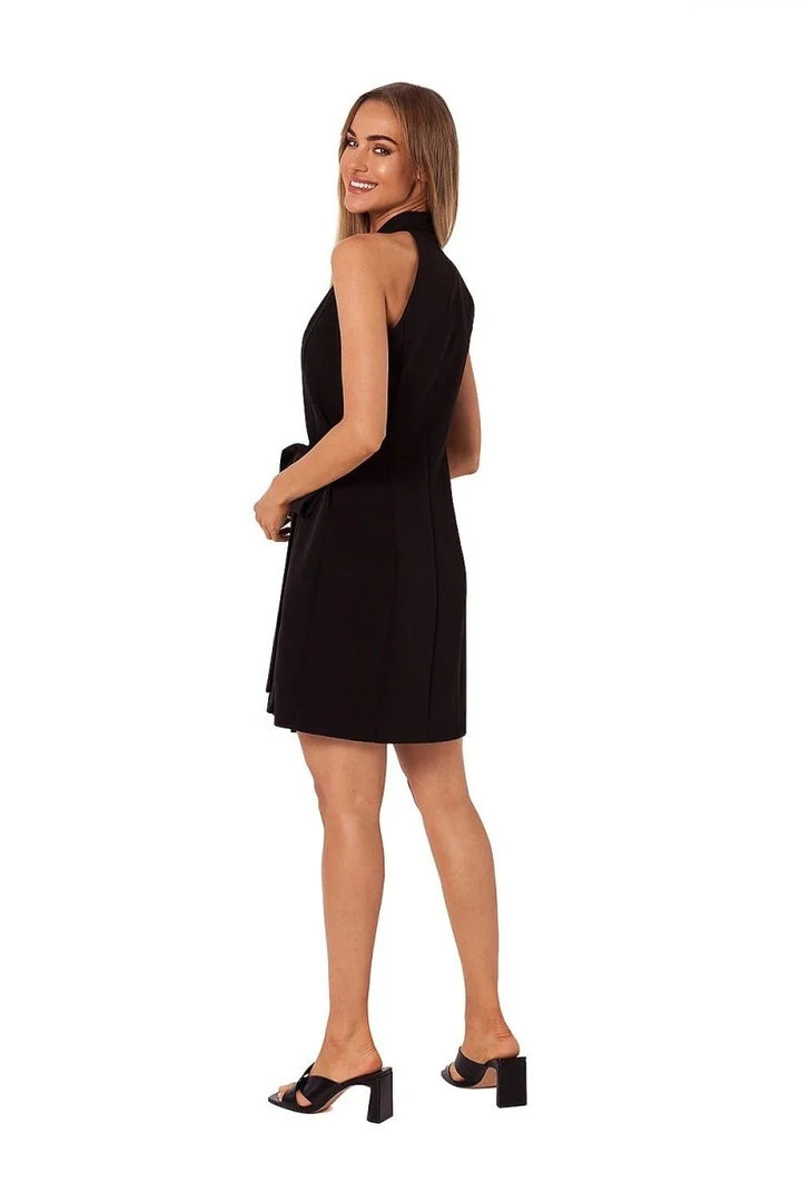 Woman in a black sleeveless dress with a tie waist, wearing black block heel mules.