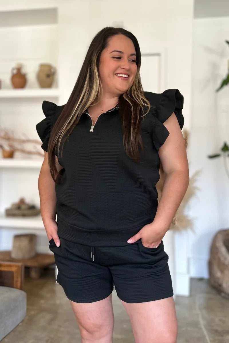Plus-size model in a black textured short suit with ruffled sleeves and a zipper neckline.