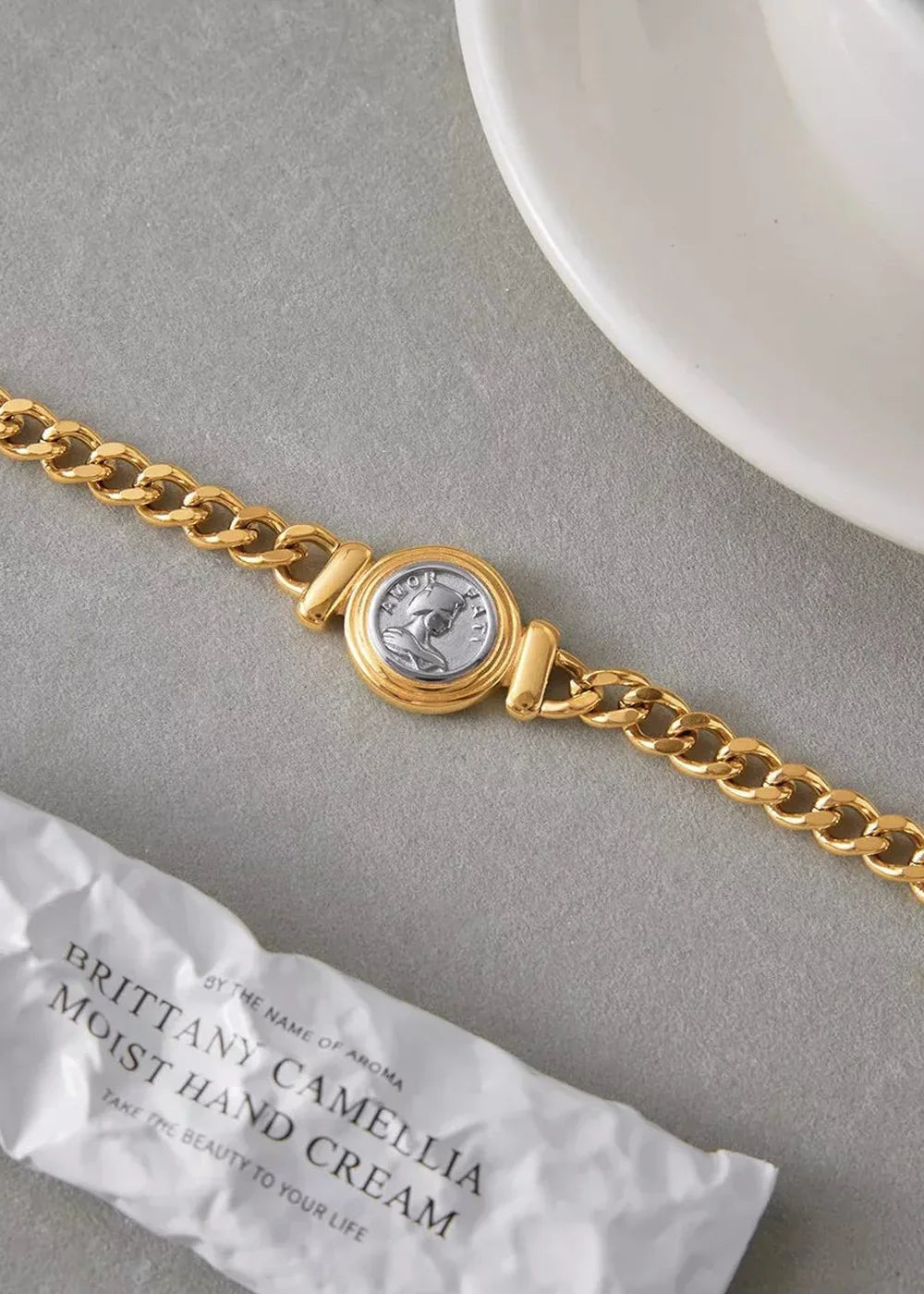 Gold chain bracelet with a silver-plated coin charm featuring a profile, resting on a grey surface next to a hand cream tube.