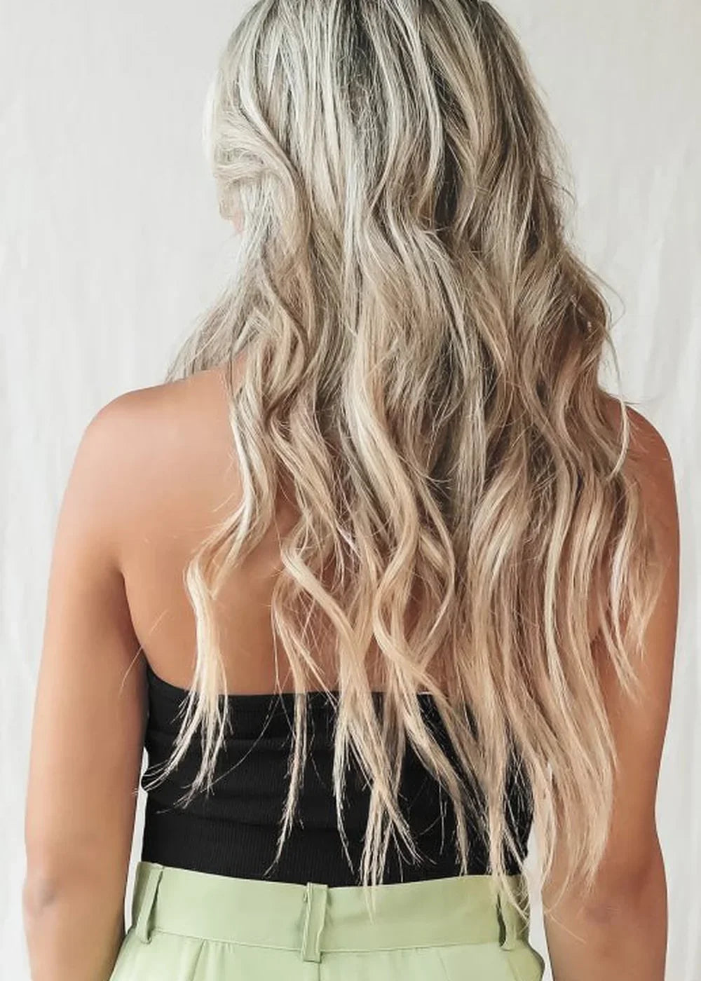 Back view of a woman with long blonde wavy hair, wearing a black strapless top and light green pants.