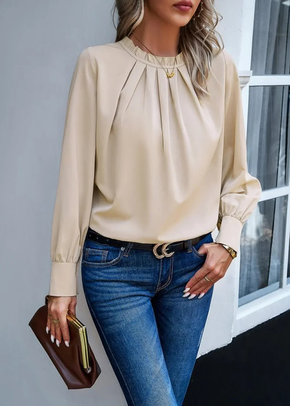 Beige long-sleeved blouse with ruffled neckline and pleated front, worn by a model with blue jeans.