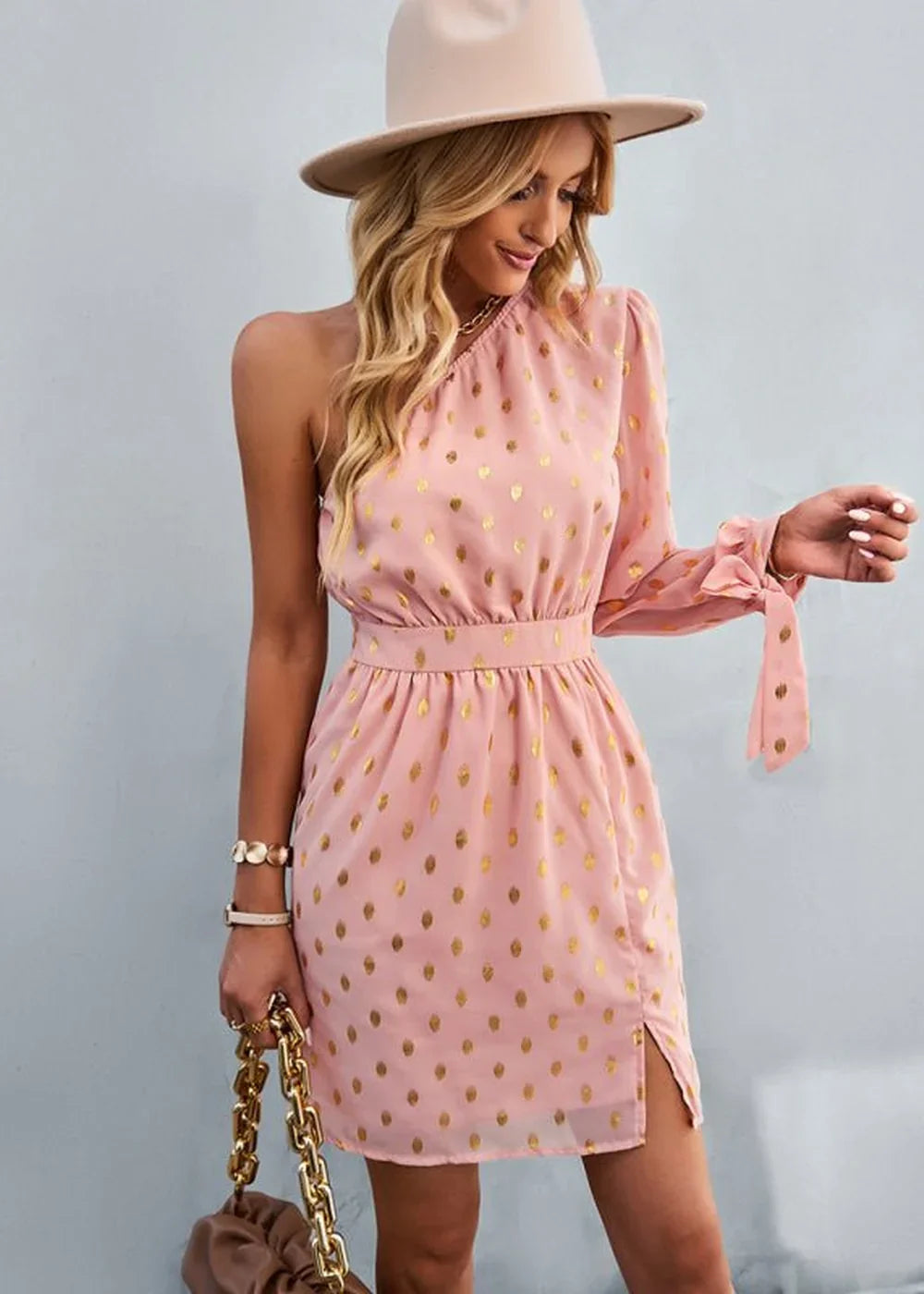 Pink one-shoulder dress with gold polka dots, cinched waist, and bow sleeve detail.