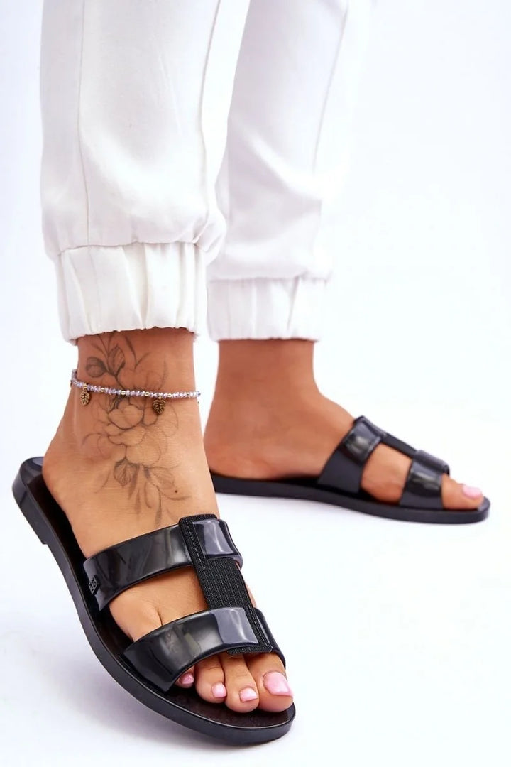 Glossy black plastic slides with a wide strap and elastic detail, worn by a model with an ankle bracelet.