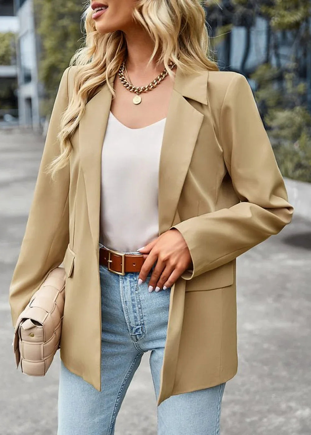 Model in a tan blazer, white top, blue jeans, and a brown belt, holding a woven clutch.