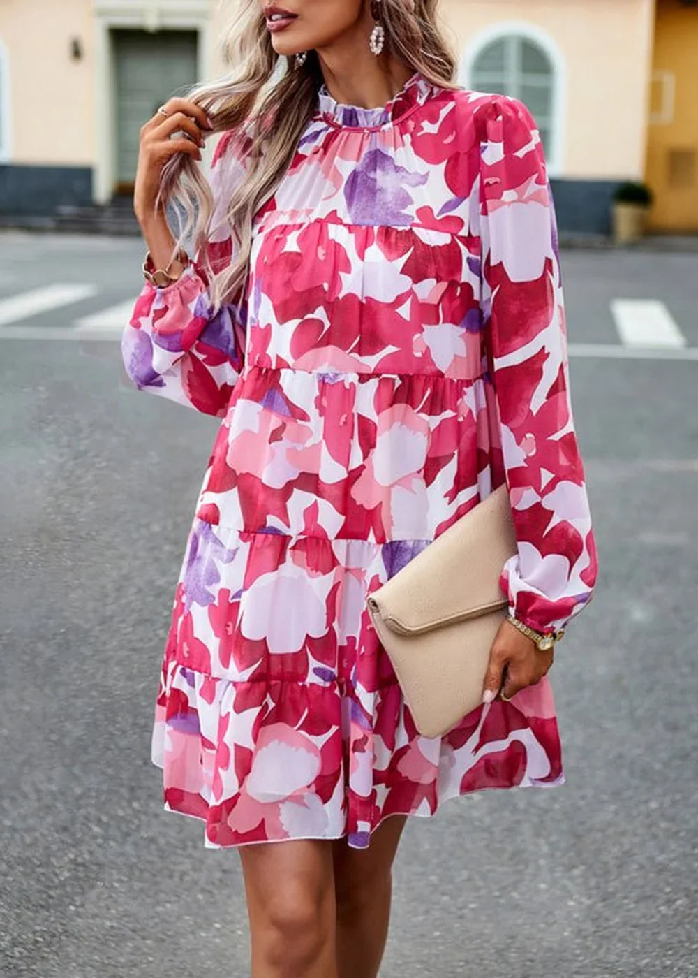Woman in a vibrant pink and purple floral print tiered mini dress with a high neckline and long sleeves, holding a beige c...