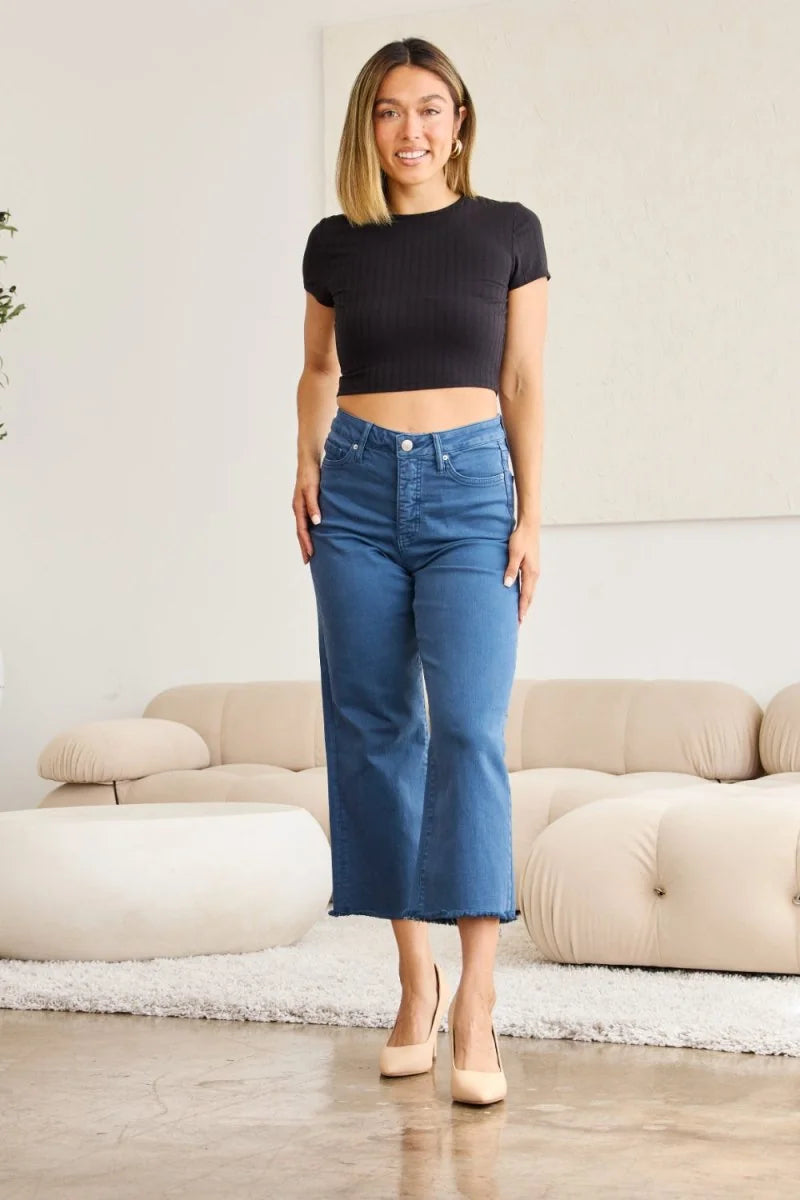 Full-length shot of a woman wearing blue denim cropped flare jeans with a frayed hem, paired with a black crop top and nud...