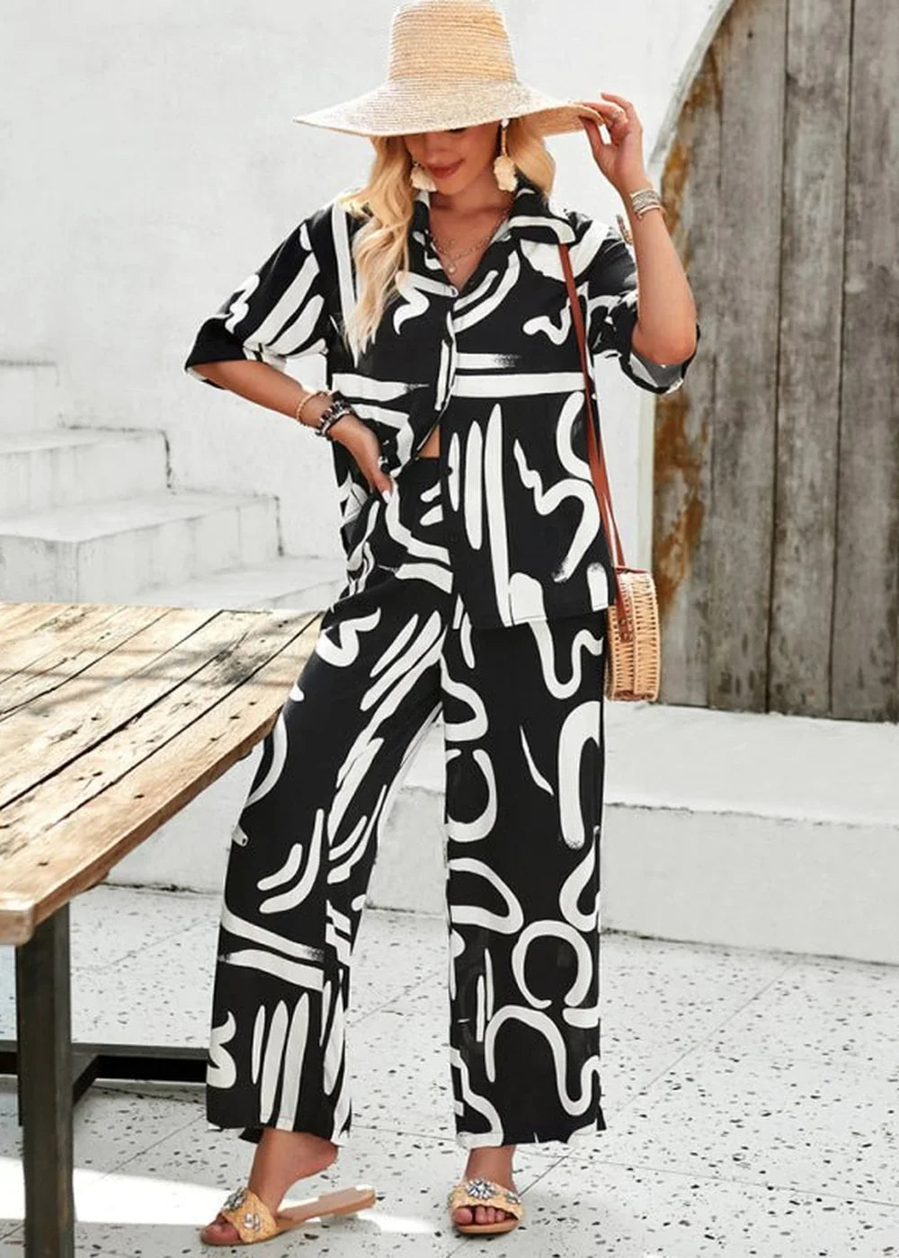 Woman wearing a black and white abstract print pant suit with a straw hat and sandals.
