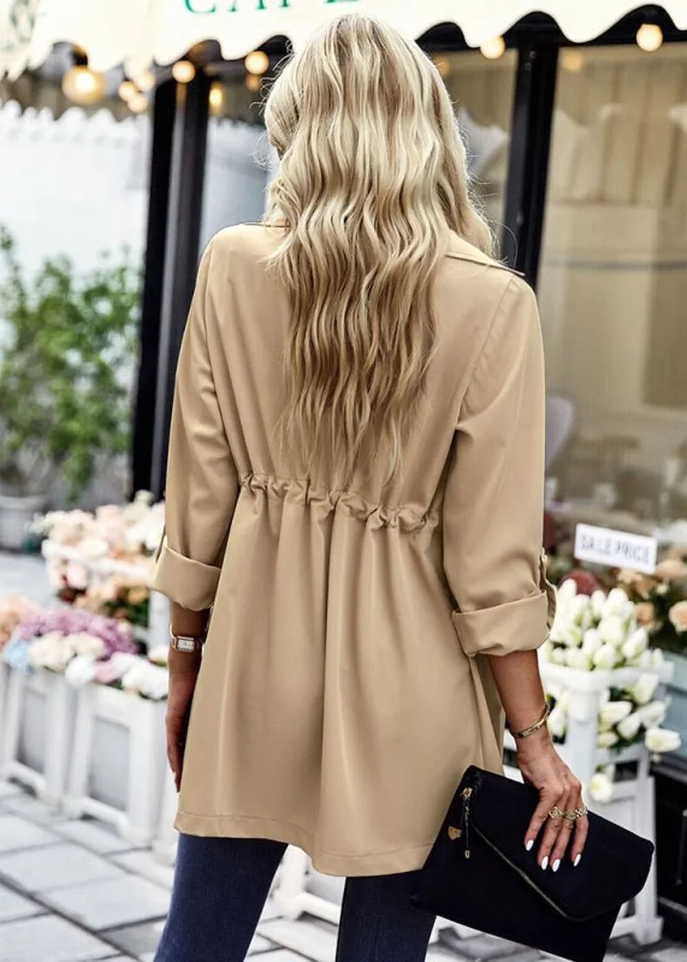 Back view of a tan trench coat with a cinched waist and rolled-up sleeves, worn by a model holding a black clutch.