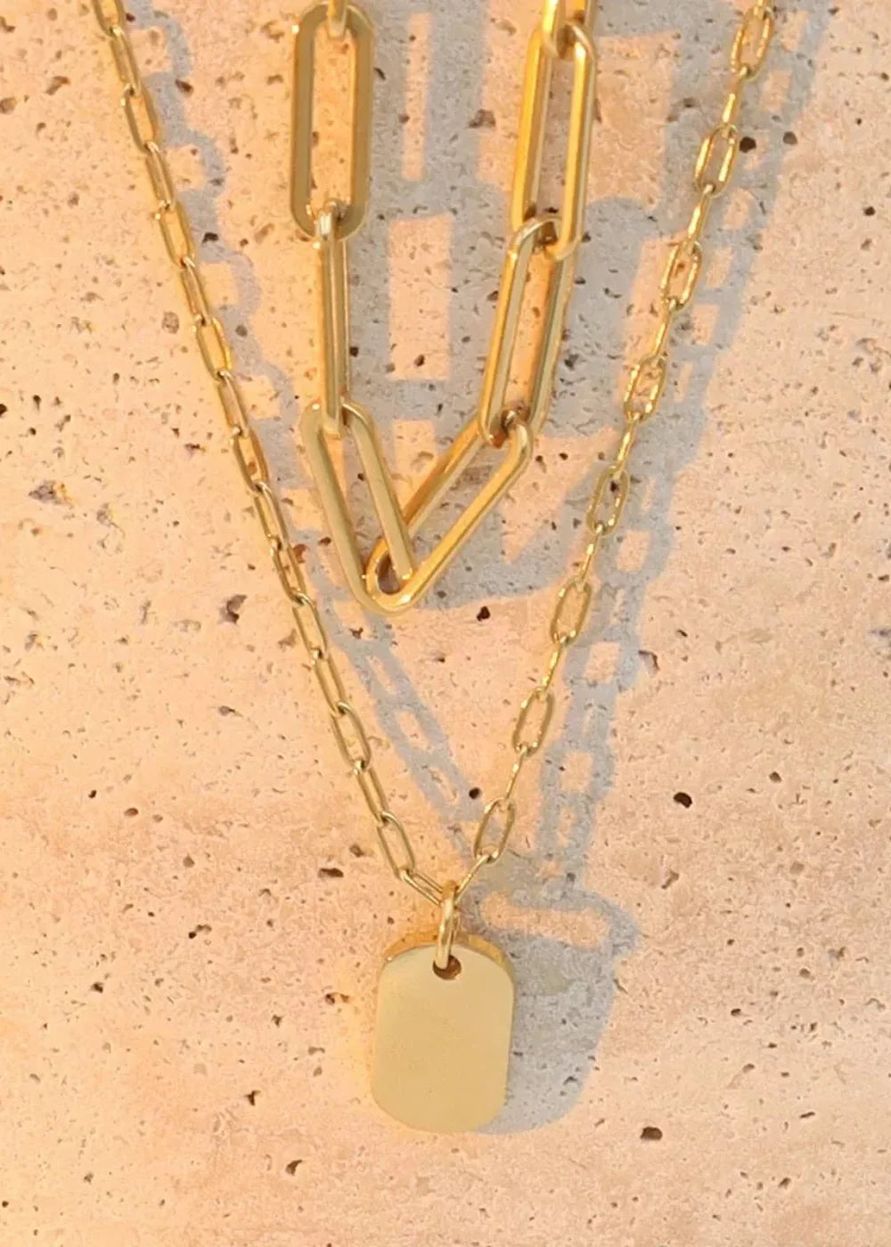 Gold-plated layered necklaces with a rectangular tag pendant on a textured wall.